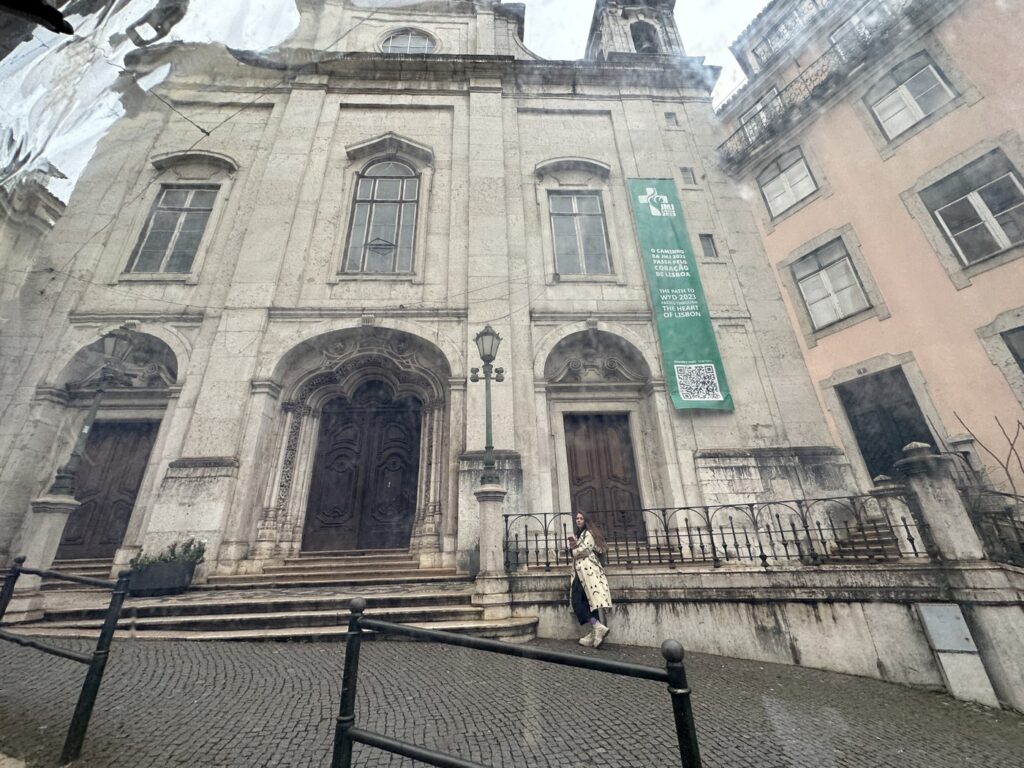 Historic stone church facade with arched doorways, tall windows, and a green banner, situated on a European cobblestone street