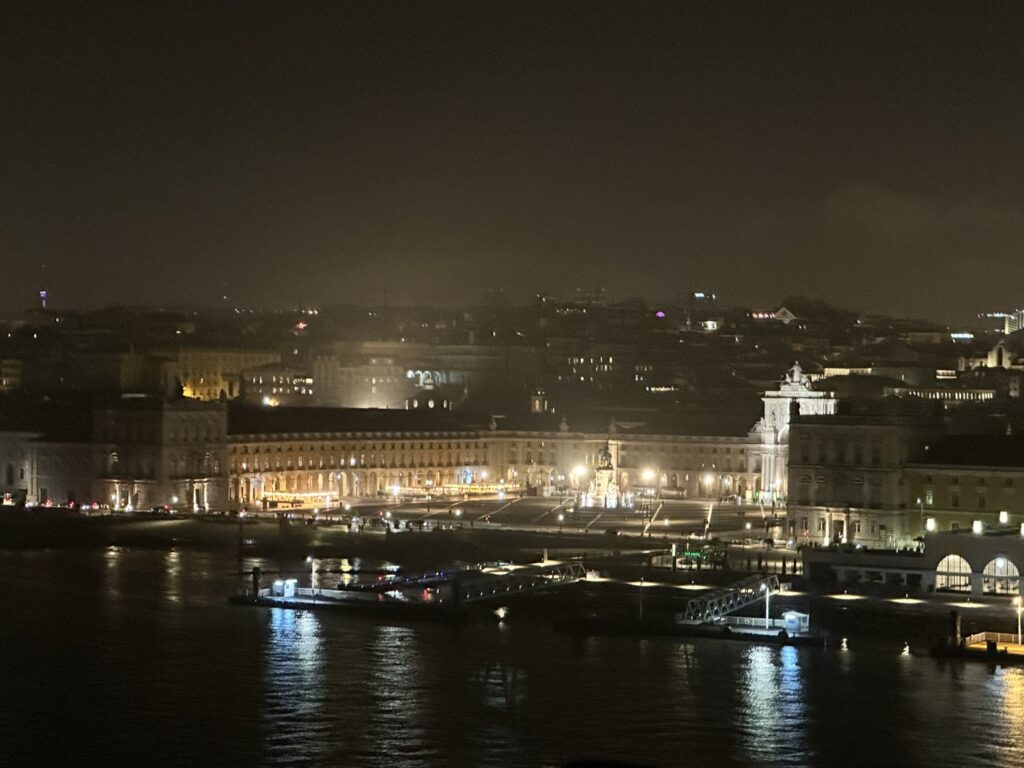 Nighttime waterfront cityscape with illuminated historic buildings, boats, and a bridge reflected in calm dark water