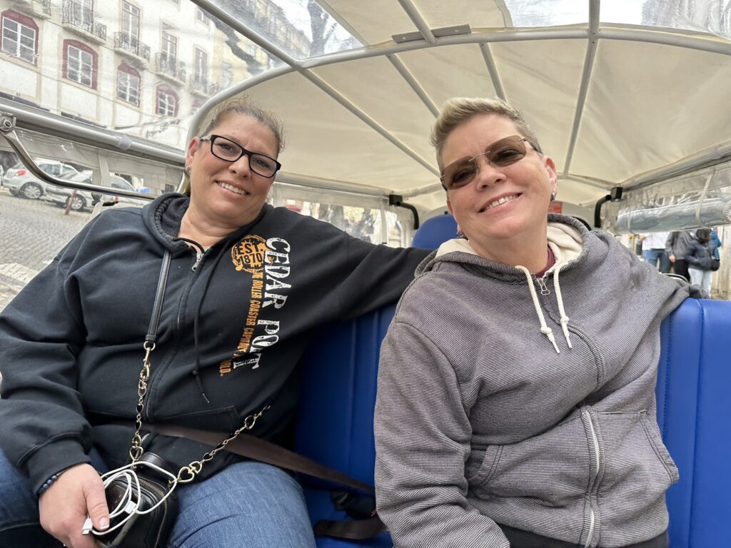 Two smiling women sit together under a transparent canopy on what appears to be public transportation in an urban setting