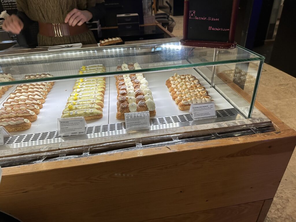 Bakery display case showcasing four rows of assorted pastries and baked goods with small descriptive labels underneath each variety
