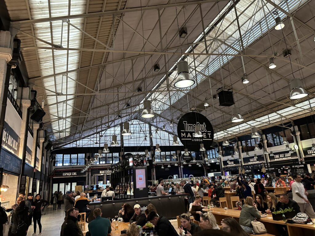 Busy indoor market hall with exposed steel ceiling, skylights, market stalls, and crowds of shoppers dining and shopping