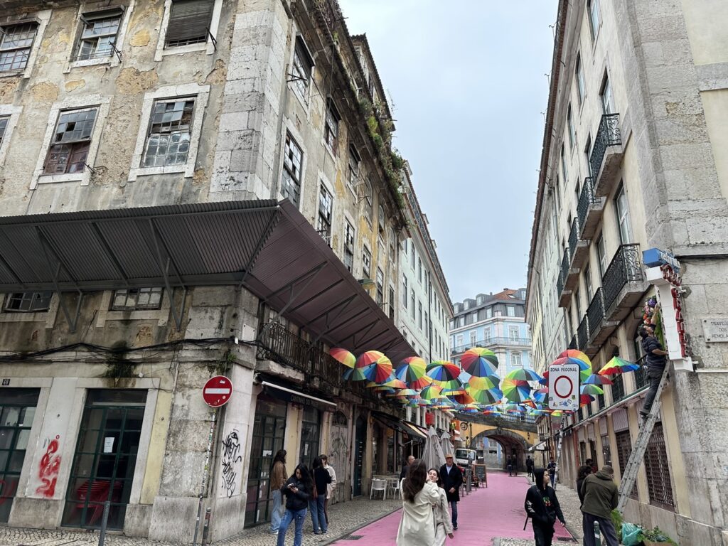 Colorful umbrellas decorate a narrow European street between historic buildings, with a pink-painted sidewalk and pedestrians walking below