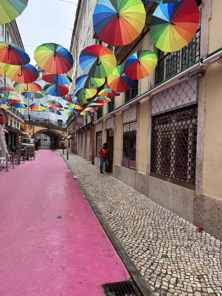 Colorful rainbow umbrellas suspended above a pink-painted pedestrian street in a European town square
