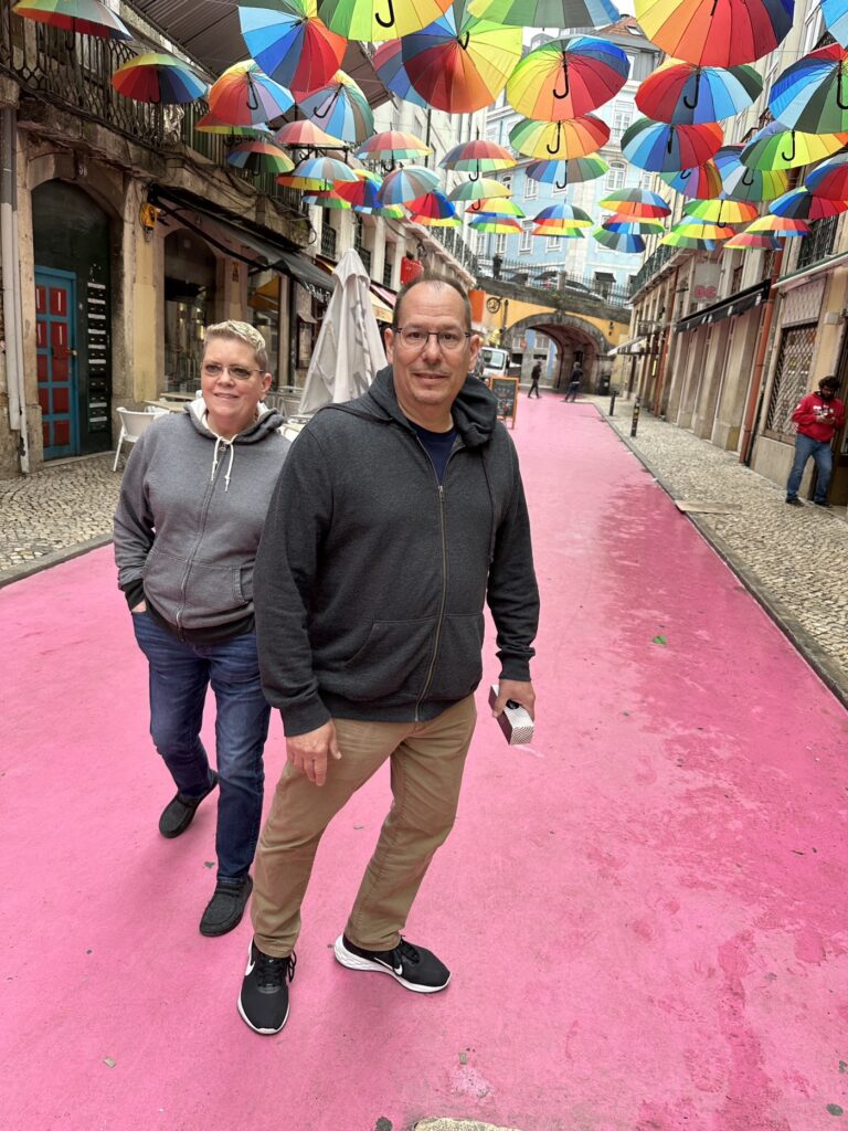 Two people walk down a pink-painted street under colorful rainbow umbrellas suspended overhead in a European town square