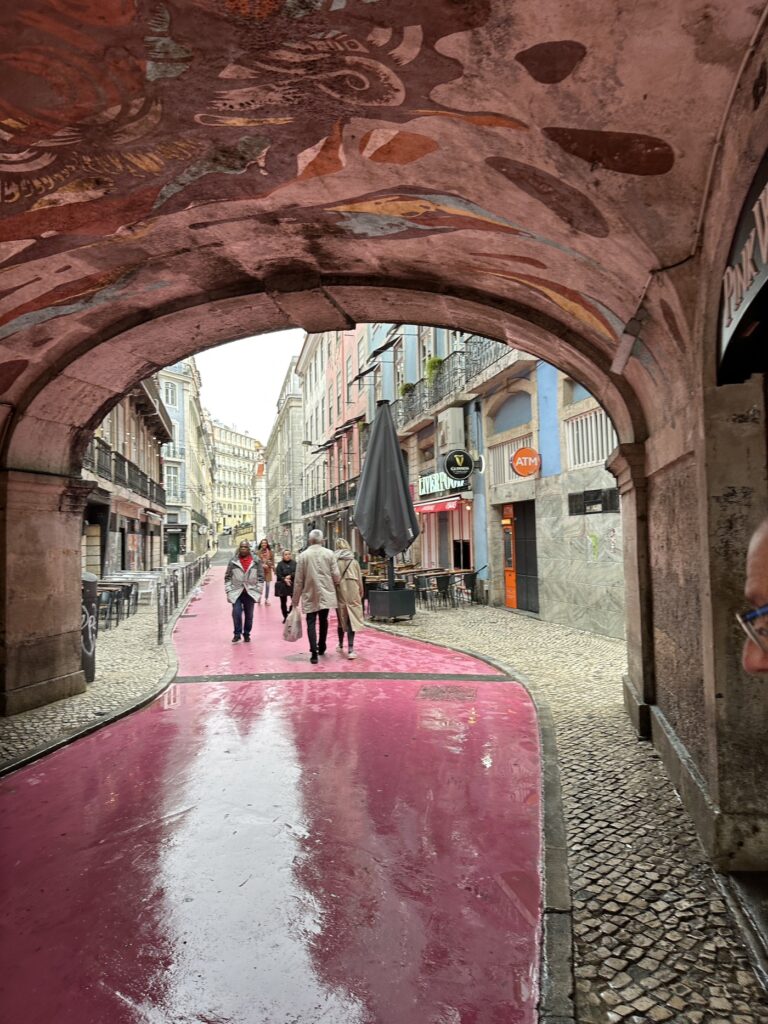 A wet pink-painted street runs through a European old town archway with historic buildings and pedestrians visible ahead