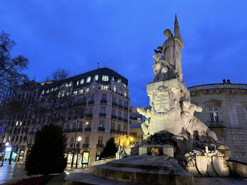 Illuminated monument statue in European city square at twilight with historic buildings and lit windows surrounding it