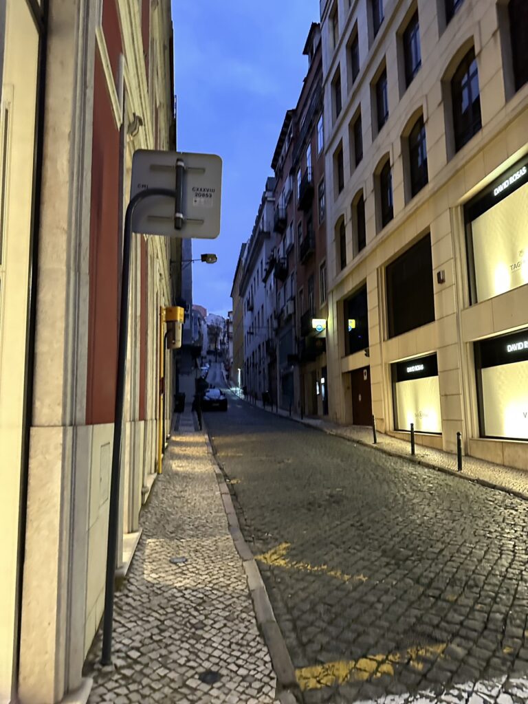 Evening street scene in a European city with cobblestone pavement, historic buildings, and illuminated storefronts lining both sides