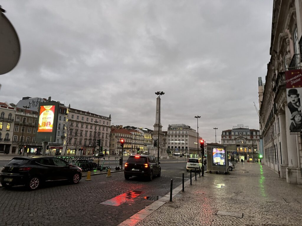 Rainy European city square with historic buildings, monuments, traffic, and wet cobblestone streets under cloudy skies