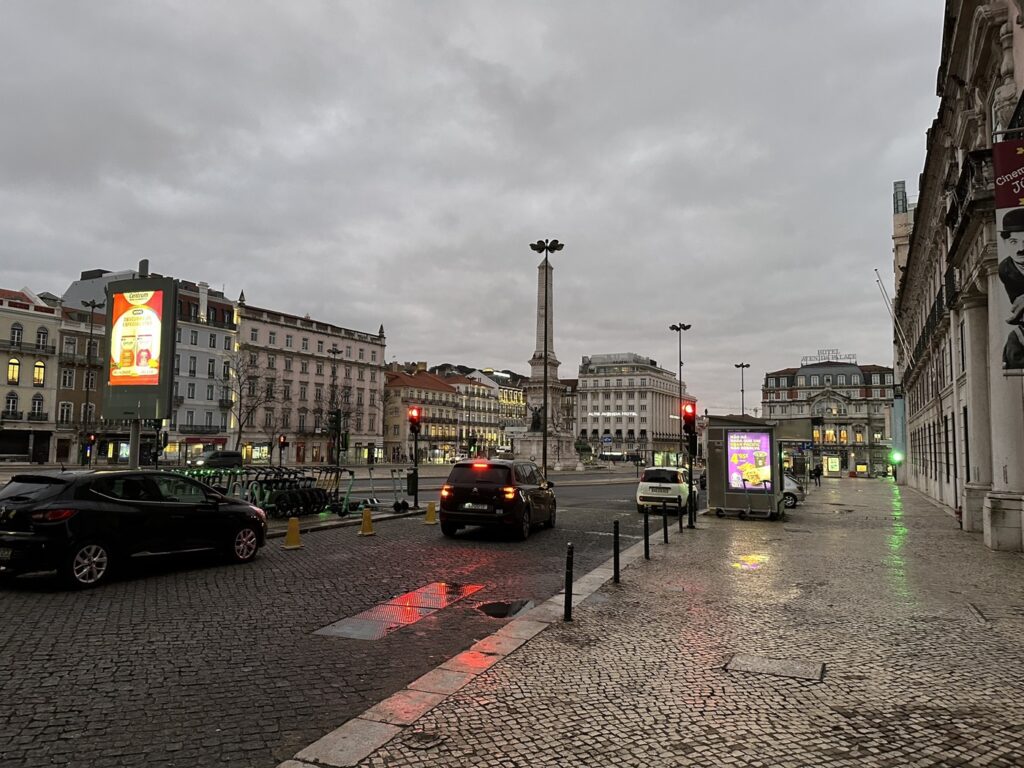 Rainy European city square with historic architecture, central monument, vehicles, and illuminated digital billboards under overcast skies