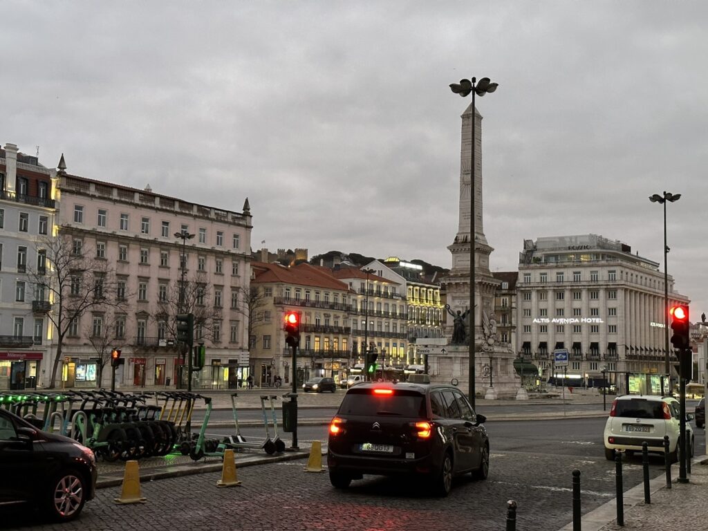 Historic European city square with central monument, classical buildings, traffic lights, and parked cars under cloudy sky