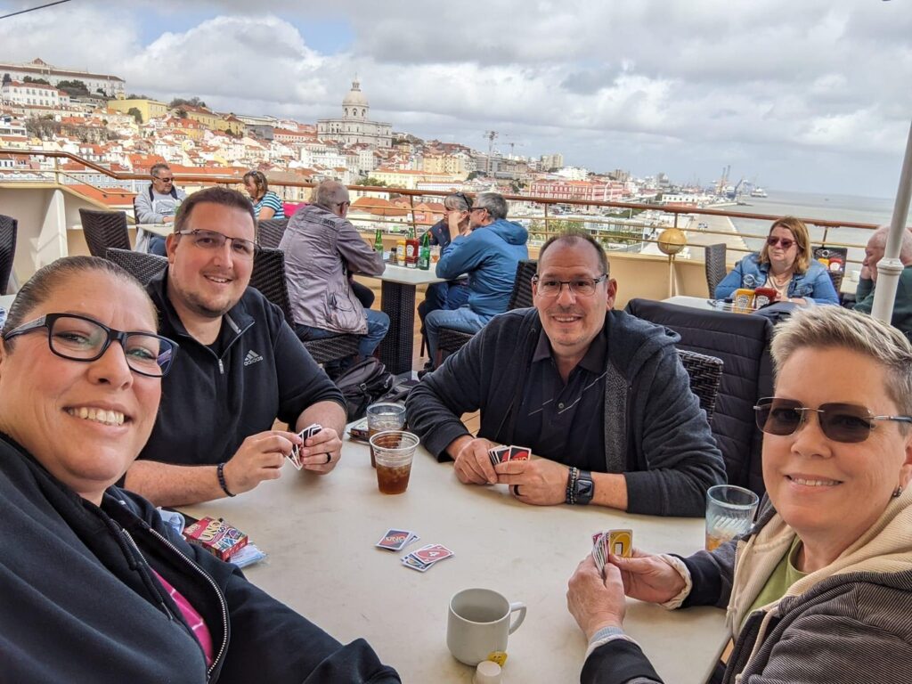Four smiling people sit at a table on a rooftop terrace overlooking a hillside European city with colorful buildings