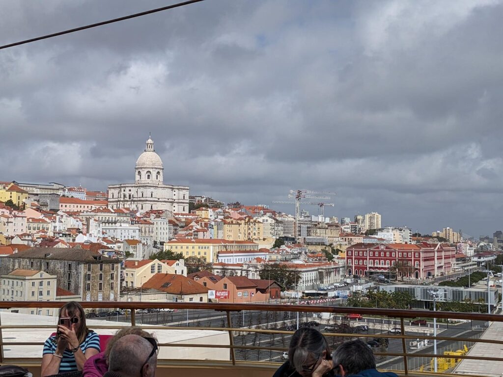 Tourists observe Lisbon's skyline featuring the white dome of the Panteão Nacional and colorful hillside buildings under cloudy skies