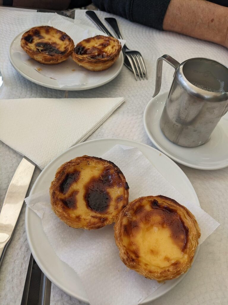 Two plates of Portuguese custard tarts with caramelized tops sit on a white tablecloth next to a metal milk pitcher and forks