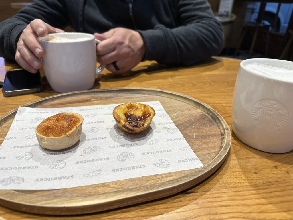 Person in dark sweater holds coffee mug at wooden table with two pastries on Starbucks napkin and white ceramic cup
