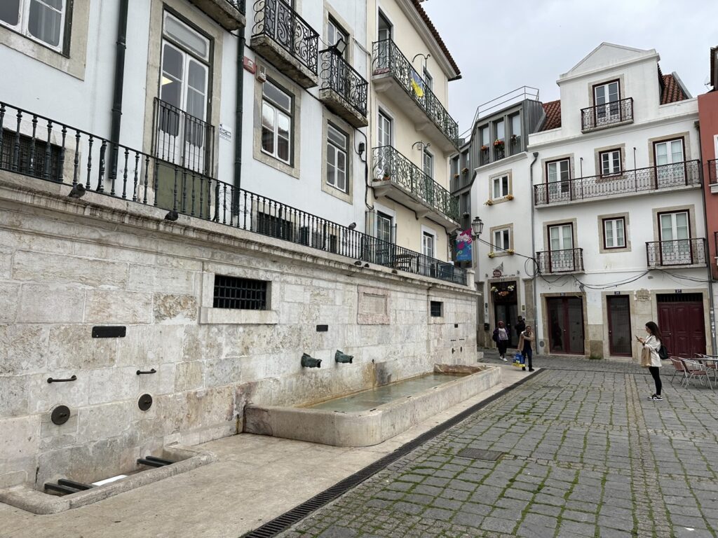 Historic European courtyard with stone buildings, ornate balconies, and a central water fountain on a cobblestone plaza