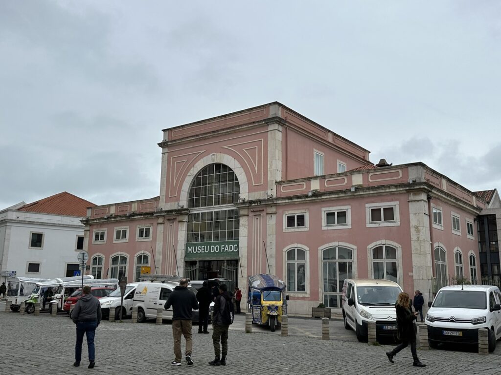 Historic pink building with "Museu do Fado" sign, featuring large arched window, surrounded by parked vehicles and pedestrians in cobblestone plaza