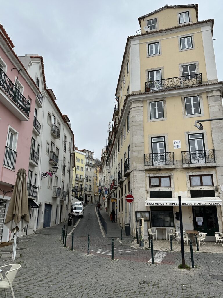 A charming European street scene with colorful historic buildings, cobblestone plaza, and ground-floor shops in what appears to be Lisbon