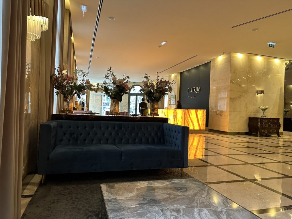 Luxury hotel lobby featuring a dark blue tufted sofa, marble flooring, decorative floral arrangements, and illuminated signage