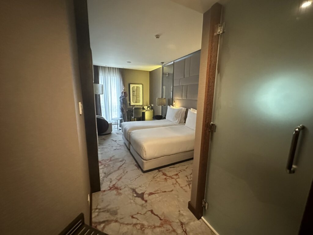 Modern hotel bedroom viewed through open doorway, featuring white bedding, elegant furnishings, and soft ambient lighting