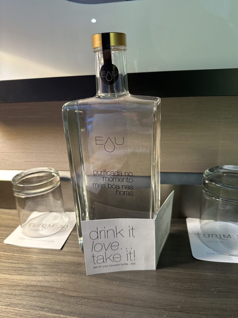 A clear glass bottle of EAU water with gold cap sits on a wooden table next to smaller glasses