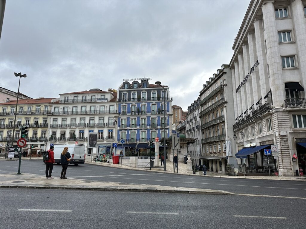 European city square with historic Haussmann-style buildings, street lamps, and pedestrians under an overcast sky